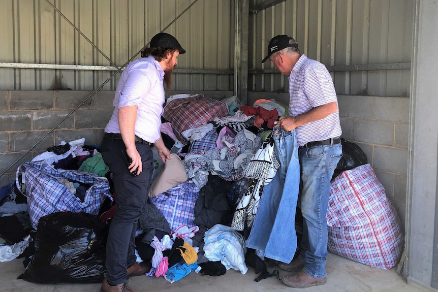 Two men standing in front of a large pile of clothing inside a corrugated iron shed.