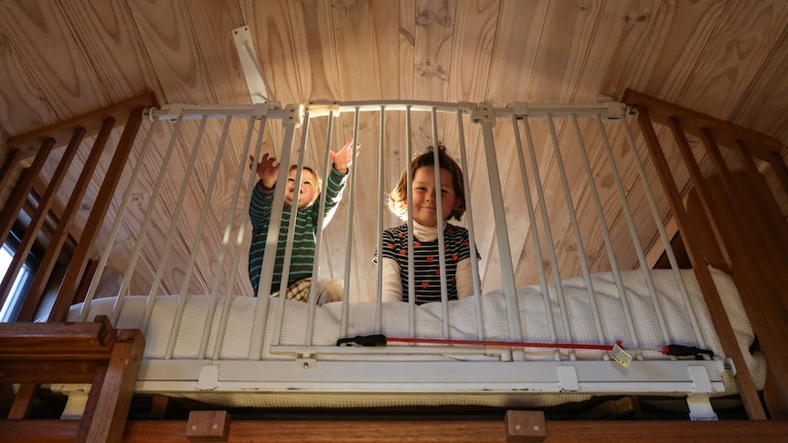 Two smiling children look out between the bars of a cot bed