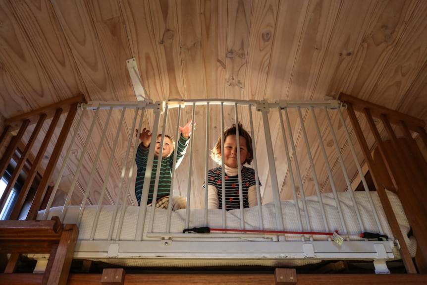 Two smiling children look out between the bars of a cot bed