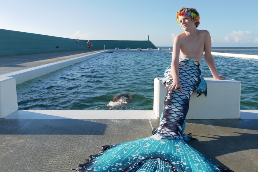 A teenage boy sits at the end of a pool block with a long blue mermaid tail and flower crown.