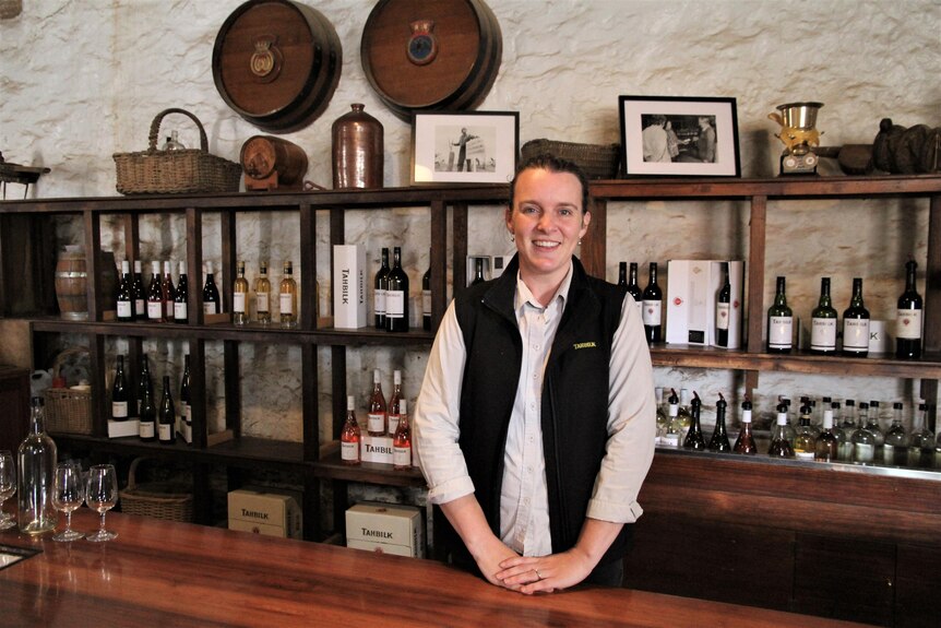 A smiling woman in a dark waistcoat stands behind a large bar.