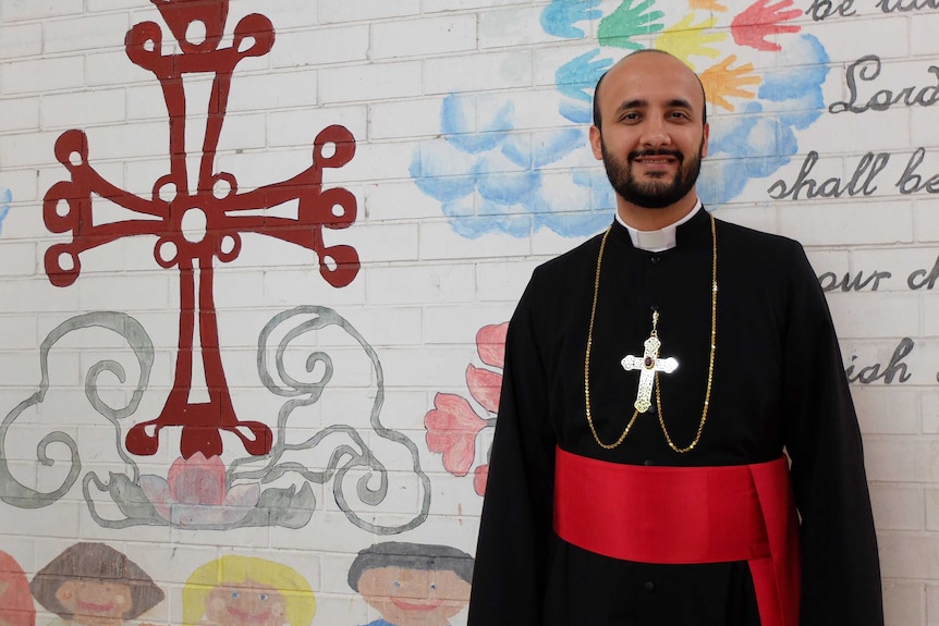 Assyrian Bishop Mar Benyamin Elya standing wearing religious robes and standing against mural wall.