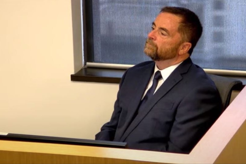 A screen shot from an internet video stream of Michael Connolly wearing a suit, sitting in a witness box giving evidence.