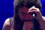 A man looks down and leans on a mic stand in tears.
