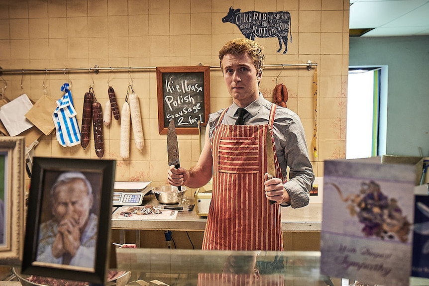A man wears a red striped apron in a butcher shop. looking at the camera, he holds a knife in front of an array of sausages.