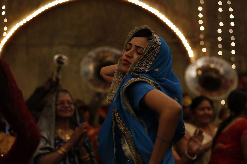 Woman in blue sari dancing with her eyes closed; older woman in the background.