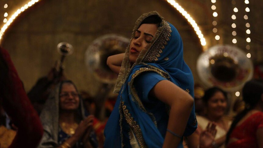 Woman in blue sari dancing with her eyes closed; older woman in the background.