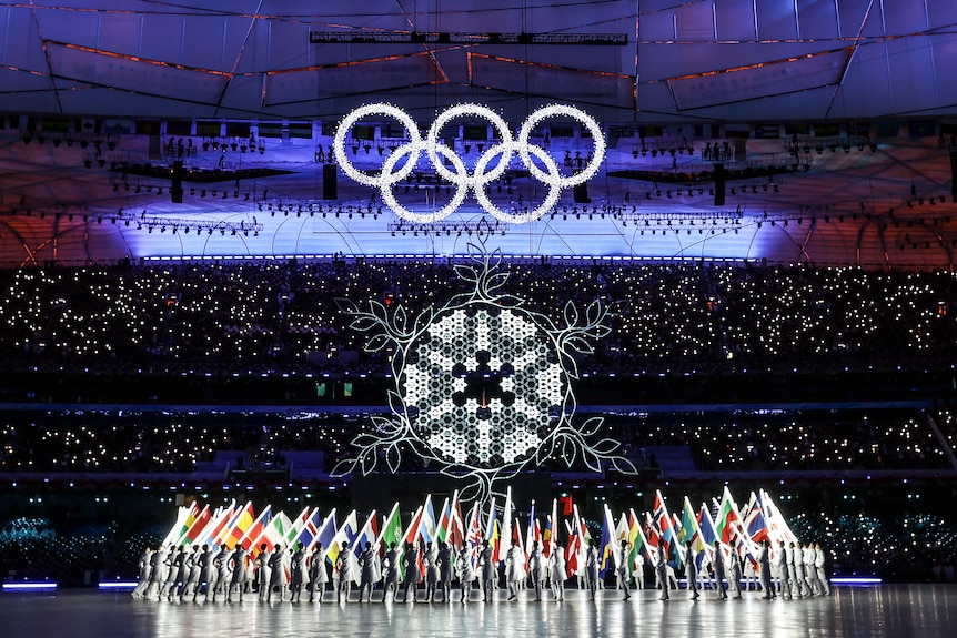The five Olympic rings hang in a stadium above a cauldron in the shape of a snowflake, with athletes and flags below that.