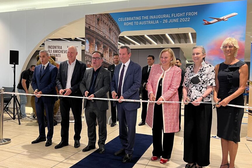 A group of seven people cut a ribbon in front of a sign that says 'Celebrating the inaugural flight from Rome to Australia'.