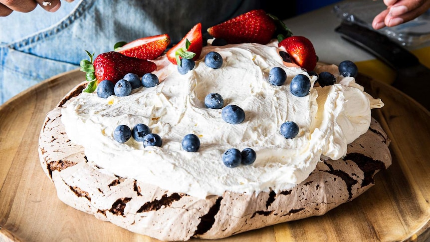 A pavlova covered in cream and berries set on a wooden platter, with hands above.