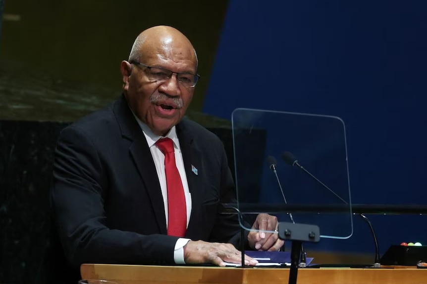 A man wearing a black suit and red tie speaking at a desk