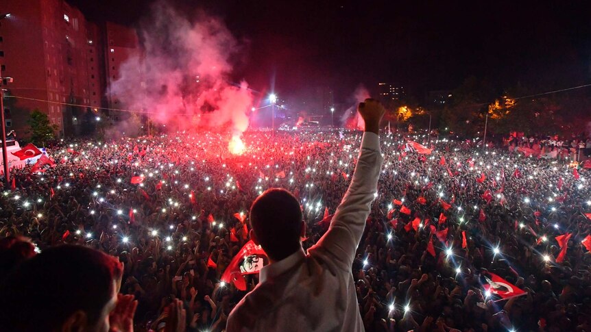 Silhouette of man in white shirt with his fist in the air as he looks at a crowd at night with red smoke coming from a flare.