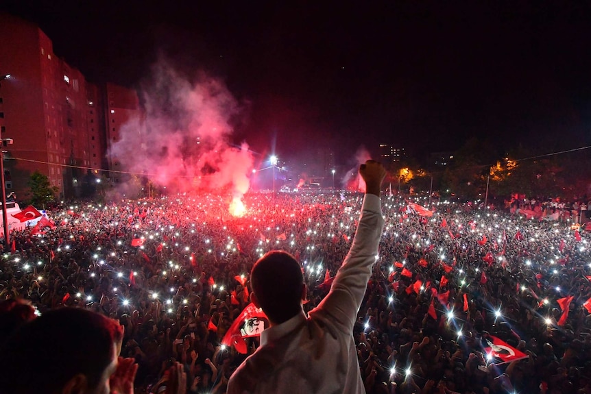 Silhouette of man in white shirt with his fist in the air as he looks at a crowd at night with red smoke coming from a flare.