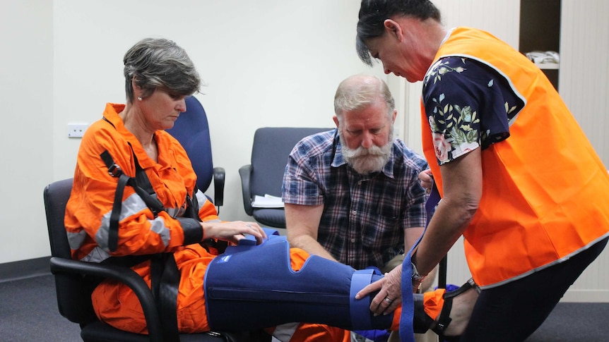 A woman sits in a chair as two other people place a brace around her leg.