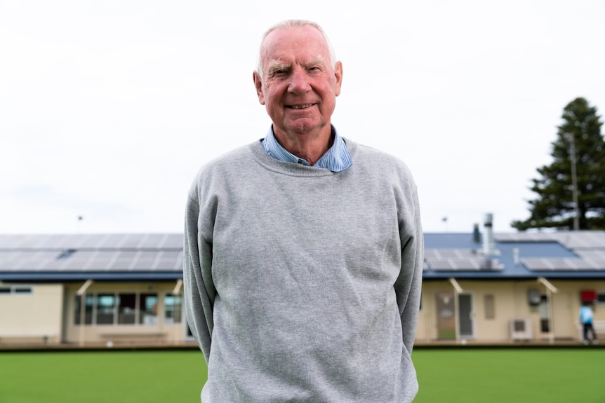 A man with grey hair wearing a grey jumper stands on a bowling green with the clubrooms behind him