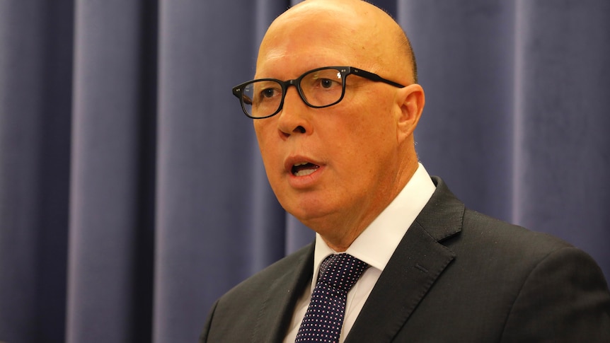 Peter Dutton stands in front of a blue curtain mid-sentence, he wears black square glasses and a suit and tie