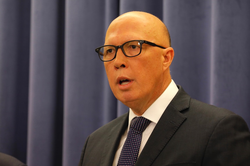 Peter Dutton stands in front of a blue curtain mid-sentence, he wears black square glasses and a suit and tie