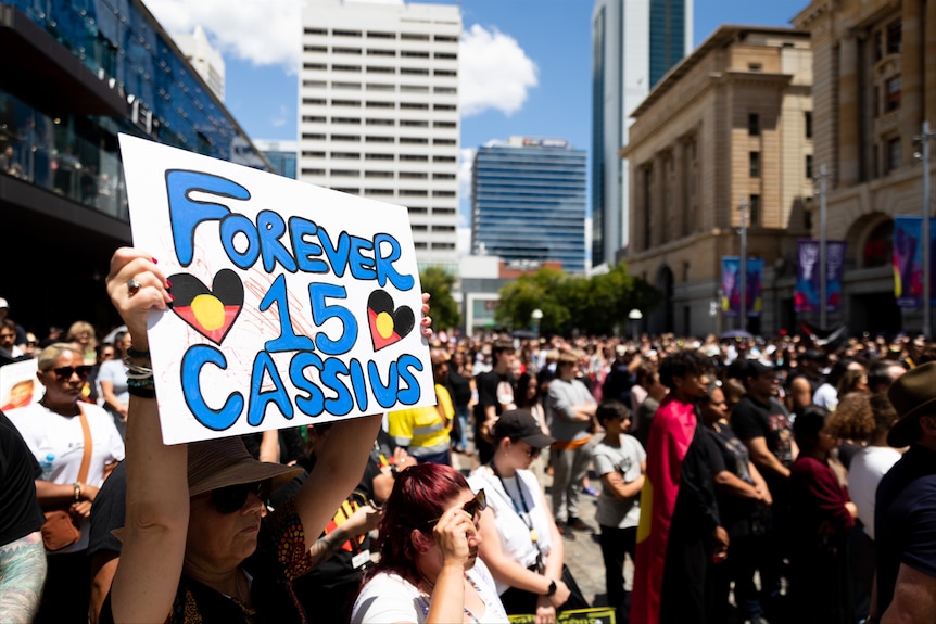 Une lecture de signe "Pour toujours 15 Cassius" est tenu au-dessus d'une foule de personnes lors d'un rassemblement à l'extérieur.