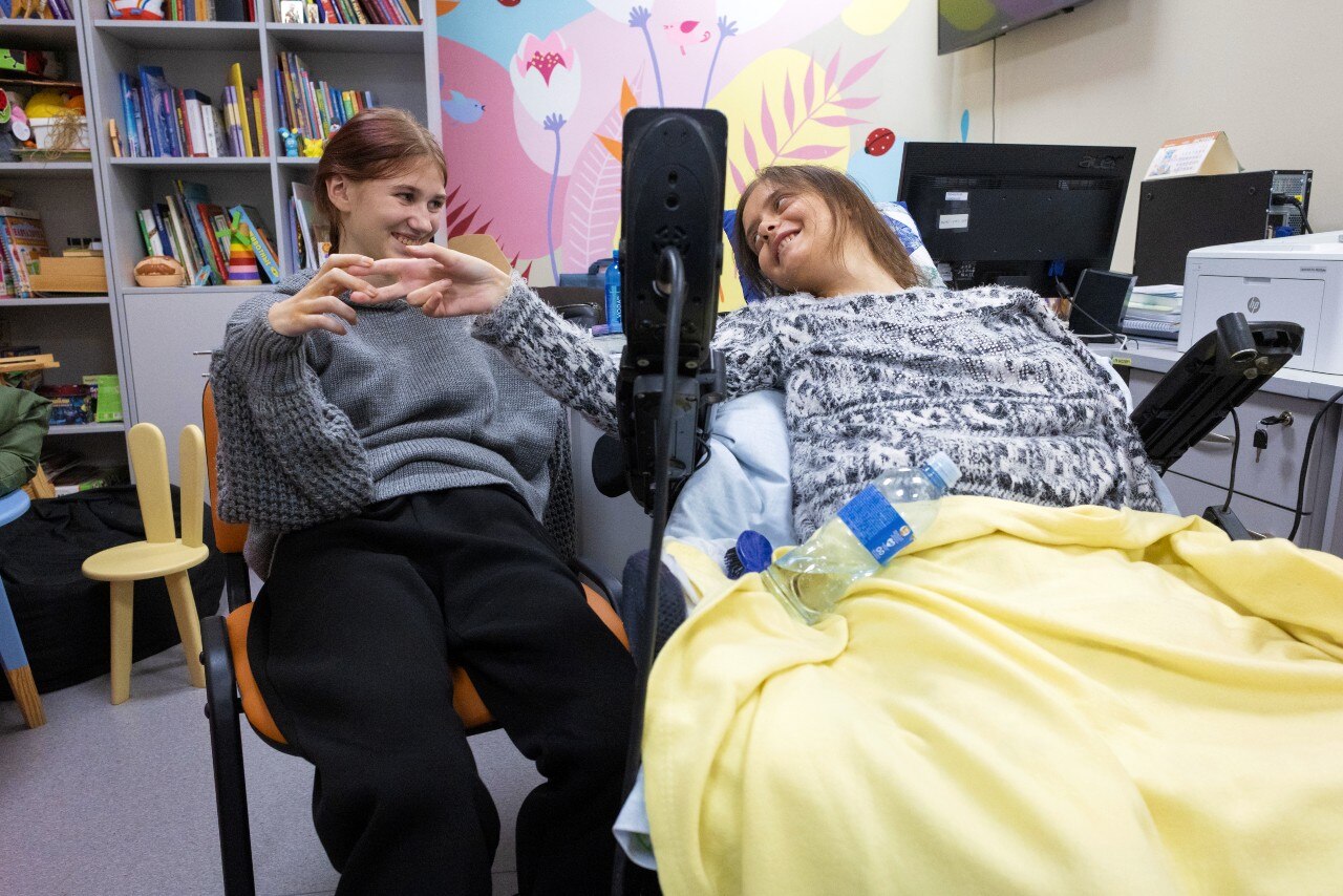 A gitl in a chair and a girl in a bed holding hands and smiling at each other.