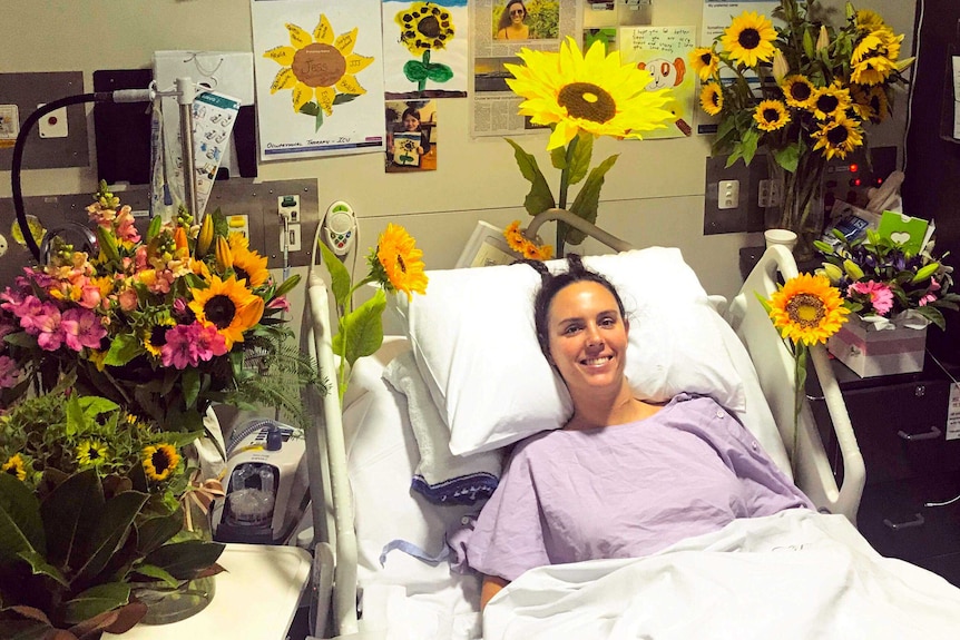 A woman in a hospital bed surrounded by sunflowers, cards, newspaper articles, photographs and paintings