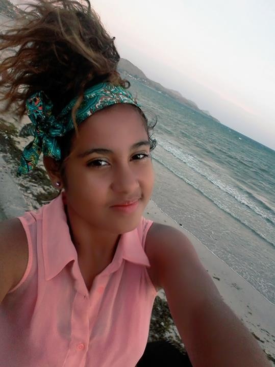 A young girl in a pink shirt and green headscarf stands on a beach