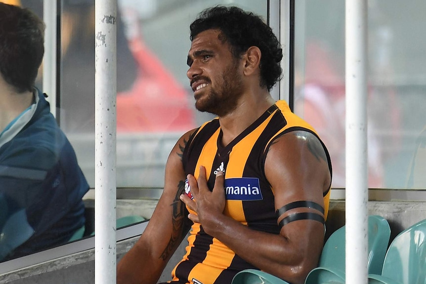 An Australian Rules player in a yellow and brown striped guernsey sits on the sidelines, clutching his chest.