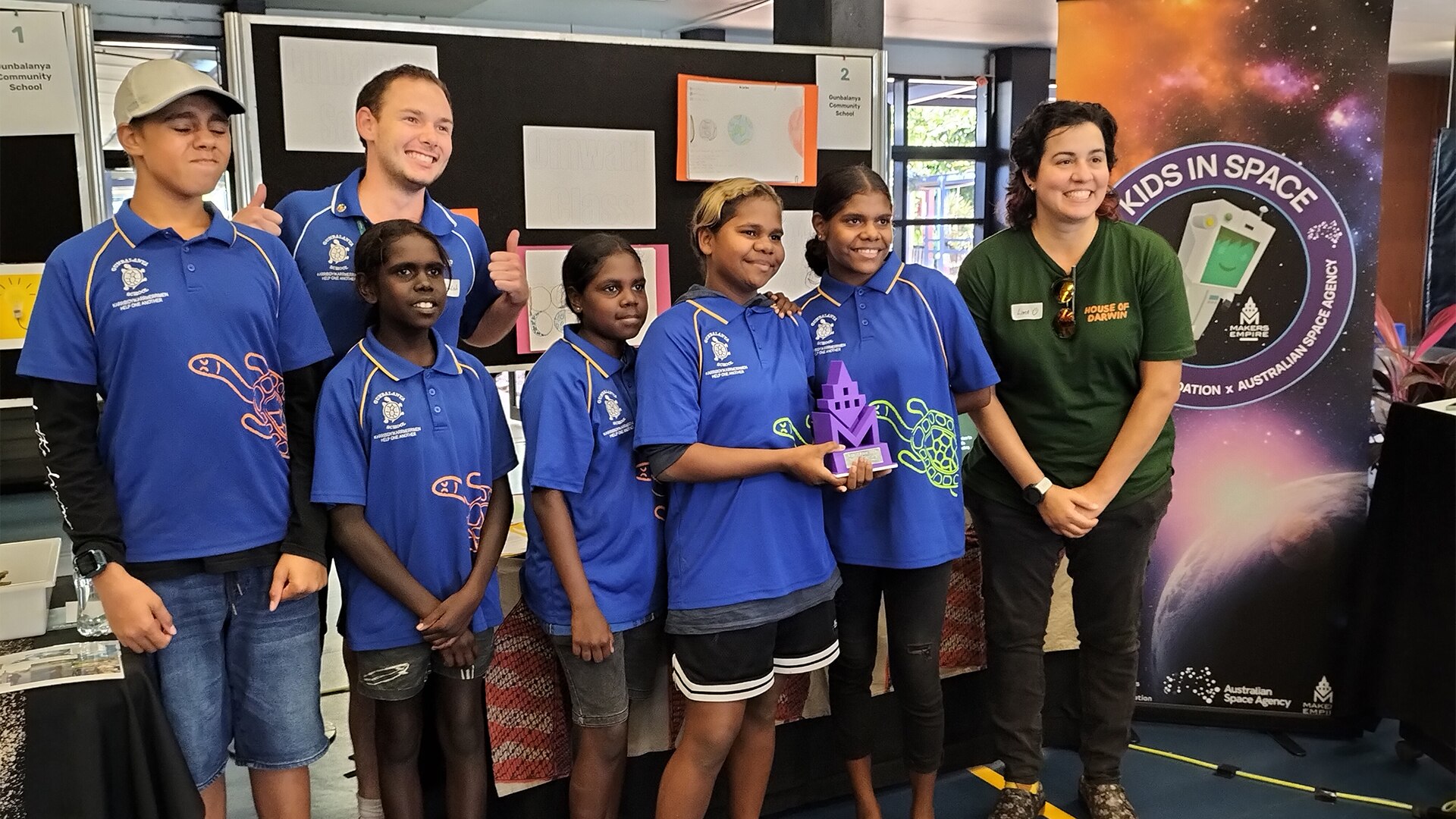 A group of students pose for a photo with an award in front of a sign that reads &#x27;Kids in Space&#x27;.