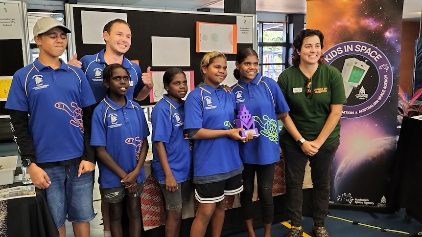 A group of students pose for a photo with an award in front of a sign that reads 'Kids in Space'.