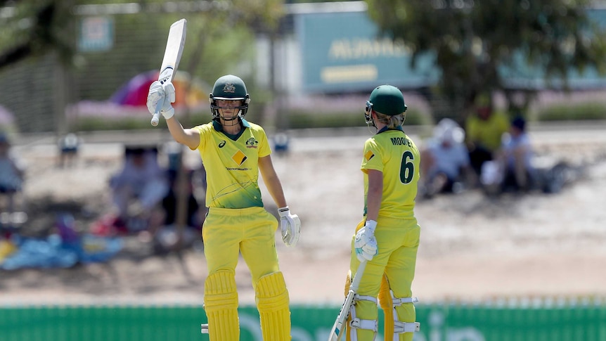 Ellyse Perry, wearing yellow cricket clothes, raises her bat in her right hand with Beth Mooney standing next to her