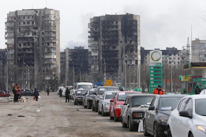 A line of cars near blocks of flats destroyed by shelling.