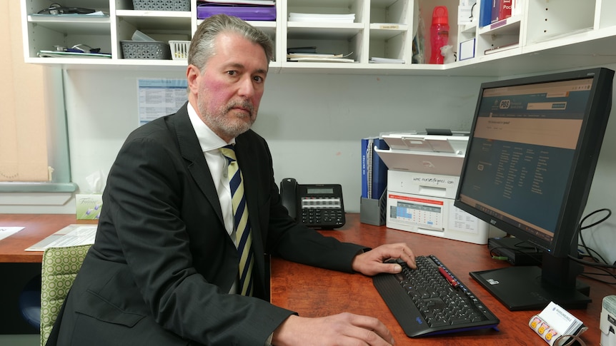 Serious man in black suit, striped tie, greying light hair, sit at desk in front of a computer, staring at camera.