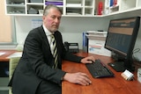 Serious man in black suit, striped tie, greying light hair, sit at desk in front of a computer, staring at camera.