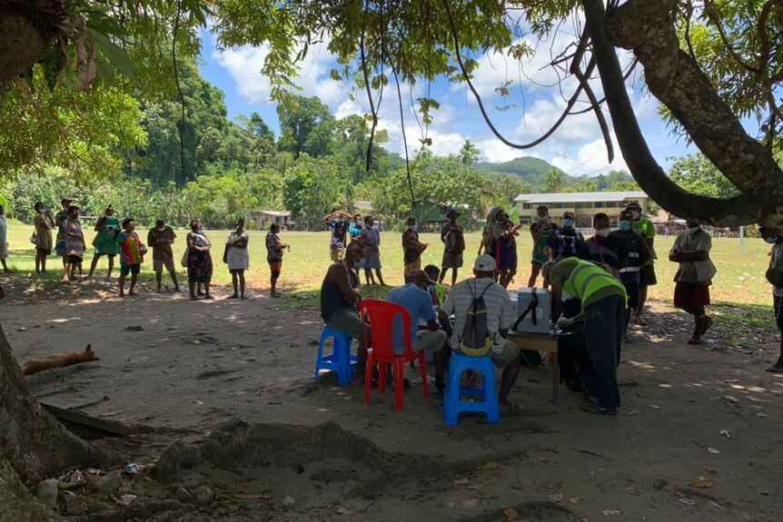 People line up under trees in front of a table.