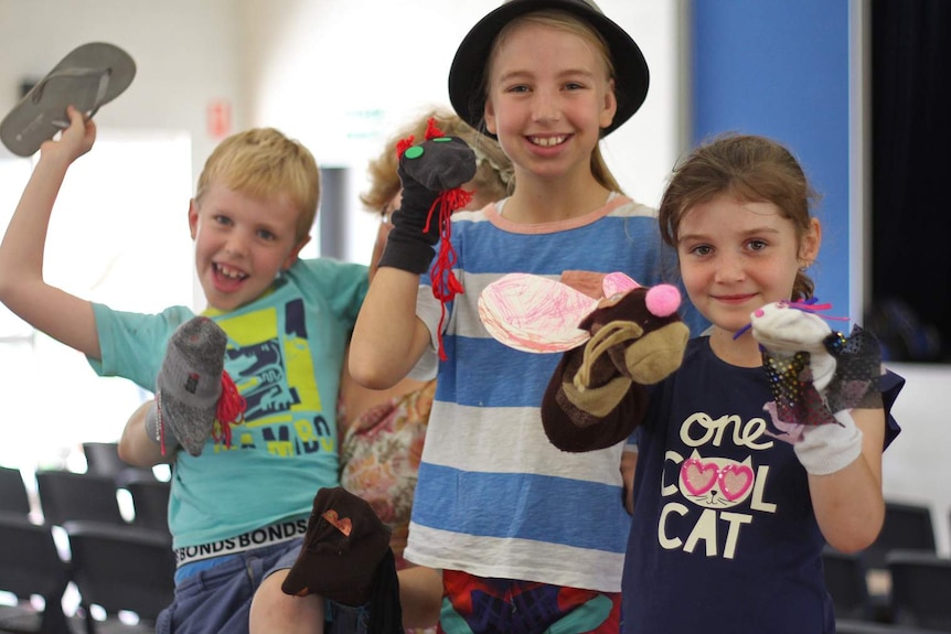 Three children wearing sock puppets smile at the camera, one has his thong held aloft