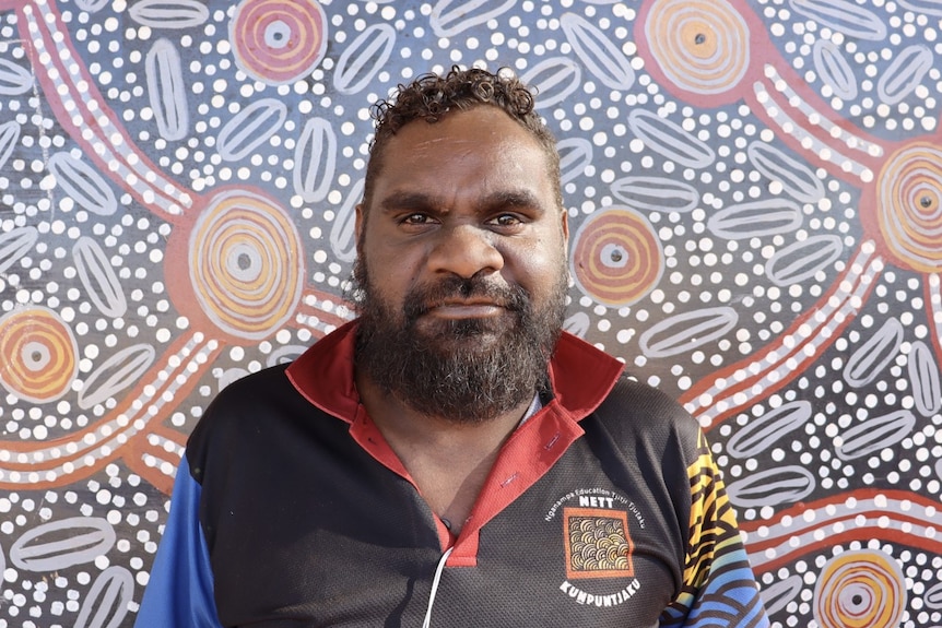 A man in front of an Indigenous painting smiles