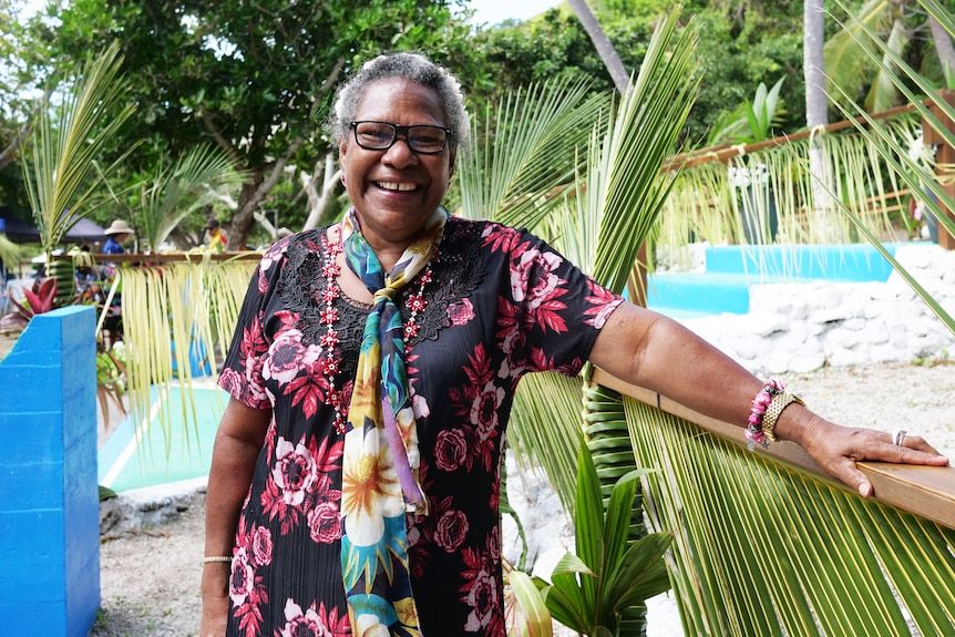 Woman in floral dress smiles at camera with arm resting on fence in front or railing decorated with coconut leaves.