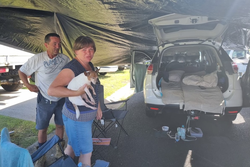 A woman and her husband stand with dog next to car boot bed under tarp tent.
