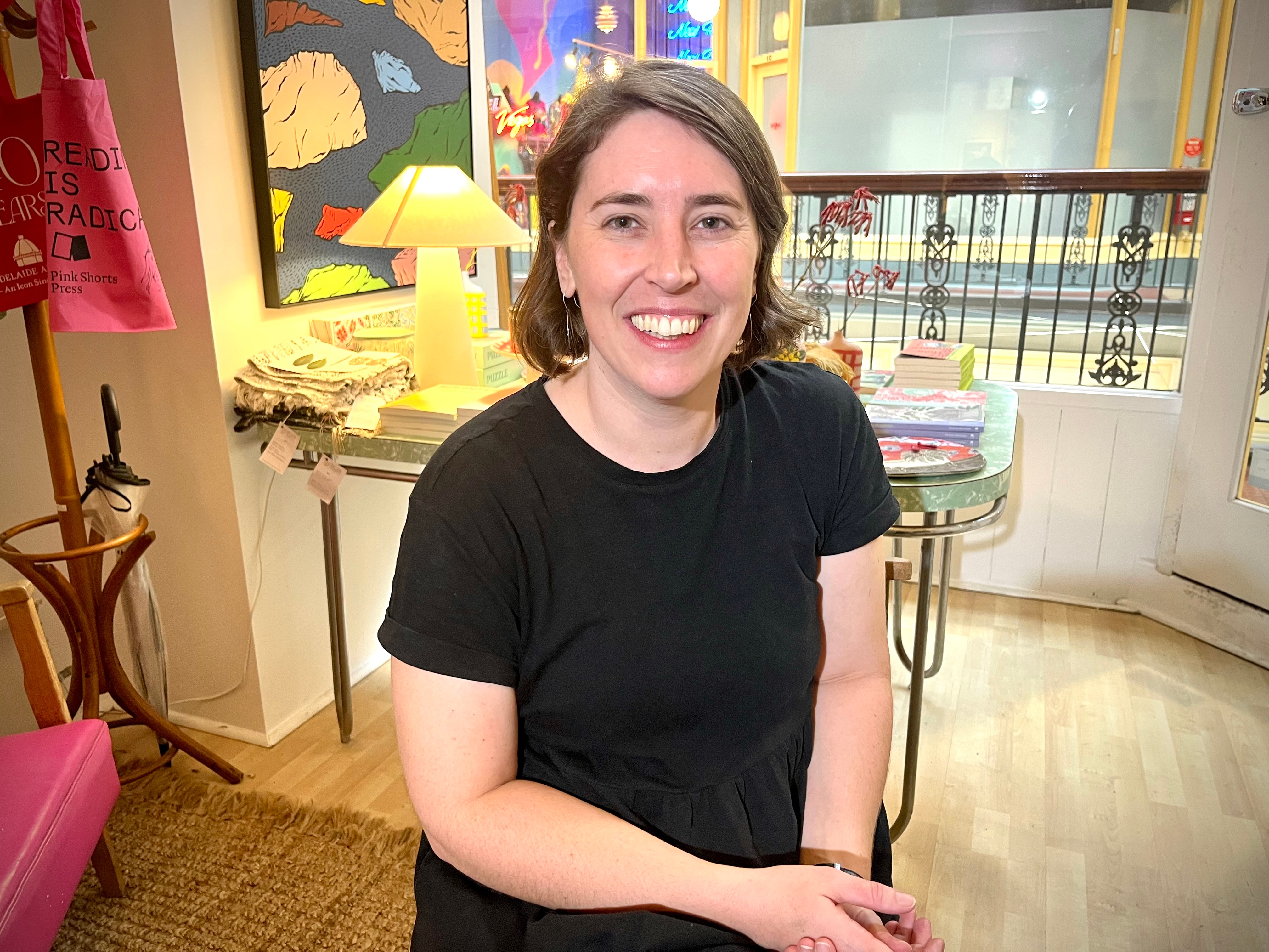 Una mujer de cabello oscuro con una camisa oscura está sentada en una habitación cálidamente iluminada y sonríe.