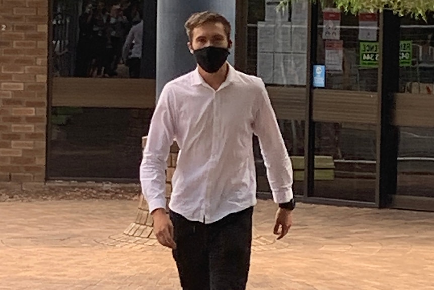 A mid shot of a young man in a white shirt and black pants walking away from a building.