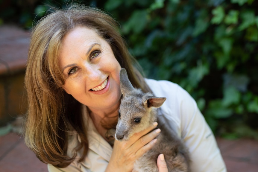middle aged woman smiles at camera holding baby kangaroo joey 