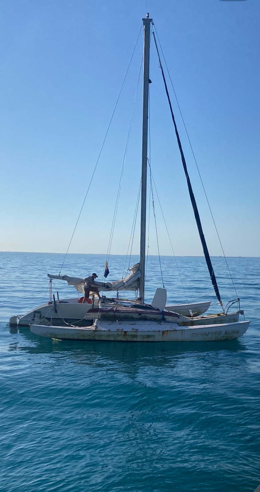 A shabby looking trimaran floating on the water with one sail wrapped at the base of the boat and a man standing on boat.