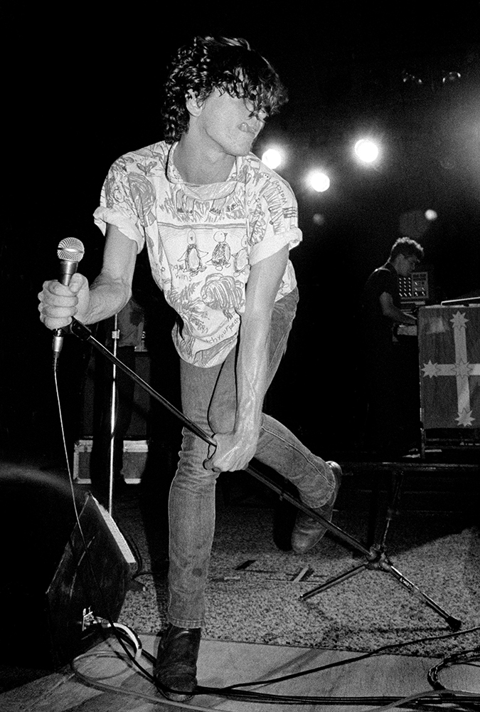 A man holds a microphone and leans forward on stage with hair covering his eyes and his tongue sticking out