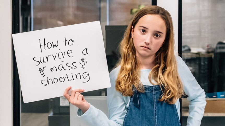 A girl in overalls holding a sign that reads "How to survive a mass shooting"