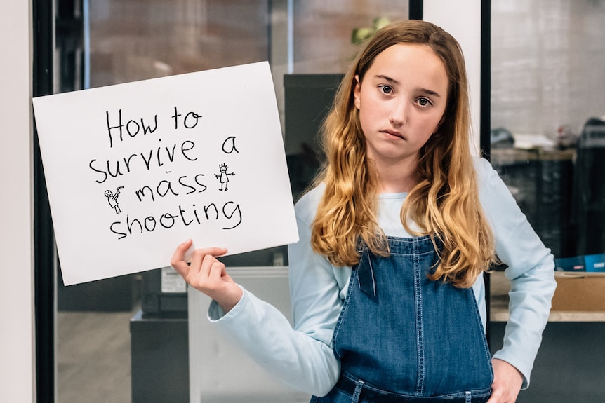 A girl in overalls holding a sign that reads "How to survive a mass shooting"