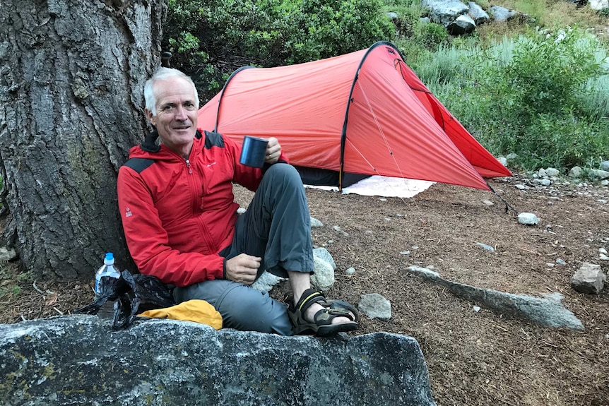 Man sits leaning against a tree drinking a cup of tea at a campsite.