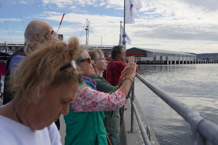 A line of people at a dock look out at the water and greet a yacht.