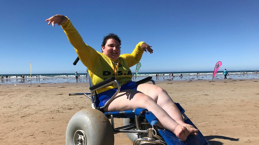 A young woman in a wheel chair on the beach. he is smiling with his hands in the air.