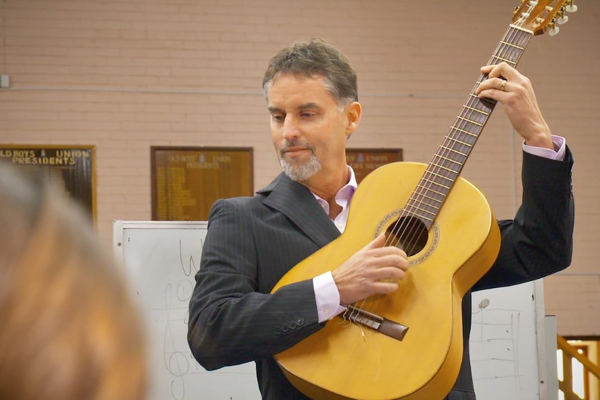 A man with a short beard instructs a class, holding a guitar high against his chest.