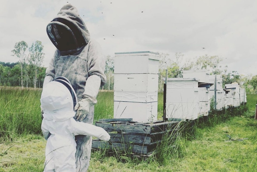 Janine Cannon and daughter Violet stand in beekeeper suits, with a row of beehives visible in the background.