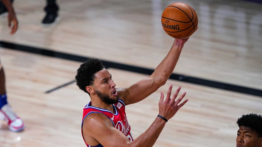 Ben Simmons holds a basketball in one hand with a grimace on his face